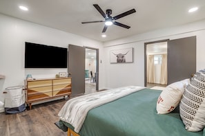 Ein Schlafzimmer mit einem Bett, einem Deckenventilator, einem Fernseher, einem Schminktisch, einem Spiegel und einer Tür zu einem weiteren Raum.