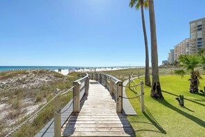 Ein hölzerner Steg führt zu einem Strand mit Palmen und einem Gebäude im Hintergrund.