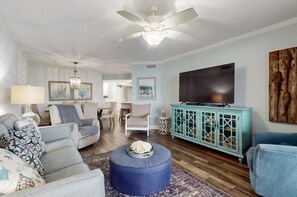 A living room with a ceiling fan, a television, a blue ottoman, and a teal cabinet. - Summer House 502B | Oceanfront | Pools, Hot Tub, Tennis | My Beach Getaways (Orange Beach)