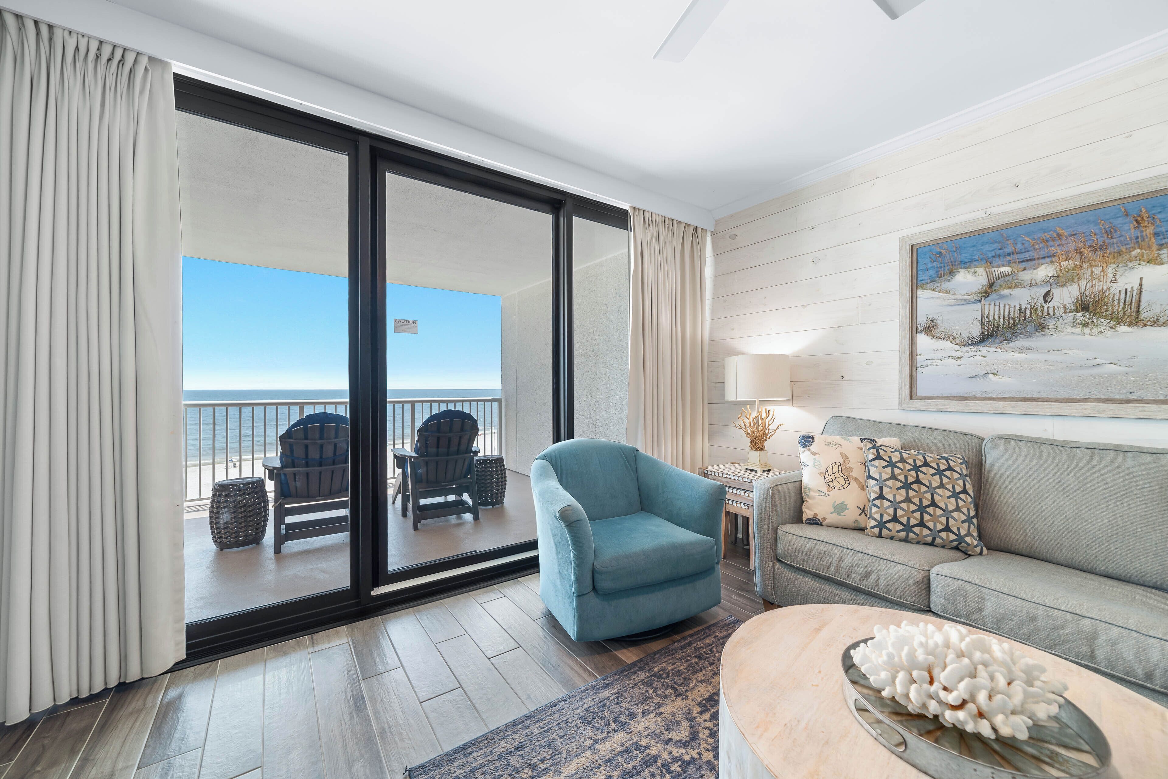 A modern living room with a view of the beach, a sofa, a blue armchair, and a coffee table.