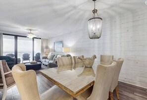 A spacious living area with a large glass-top table, multiple chairs, a ceiling fan, and a chandelier. - Summer House 502B | Oceanfront | Pools, Hot Tub, Tennis | My Beach Getaways (Orange Beach)
