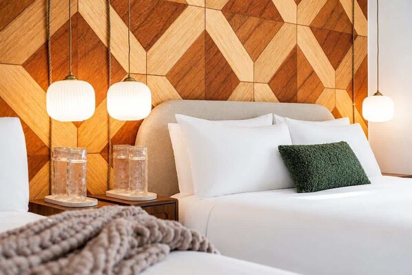 A hotel room with a geometric patterned wall, a bed with white bedding and a green pillow, two bedside tables with clear glassware, and hanging pendant lights.