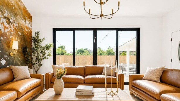 A modern living room with a large window, a brown leather sofa, a wooden coffee table, and a chandelier.