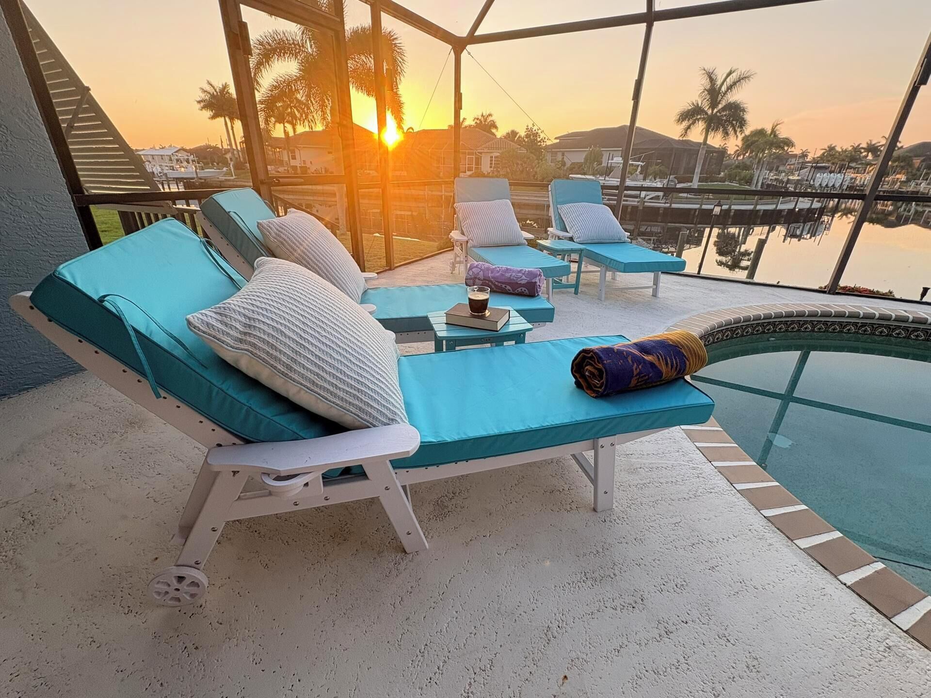 Zona junto a la piscina con sillones de descanso y una pequeña mesa, con vista a un cuerpo de agua y palmeras.