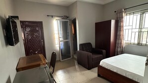 A bedroom with a bed, a desk, a chair, a wardrobe, a ceiling fan, and a window with curtains. - ESTHEMERS ROYAL HOTEL (Kumasi)