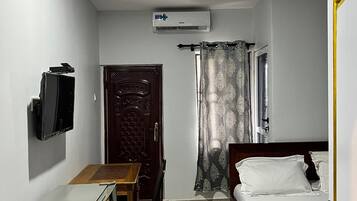 A compact hotel room with a bed, a desk, a television, an air conditioning unit, and a ceiling fan.