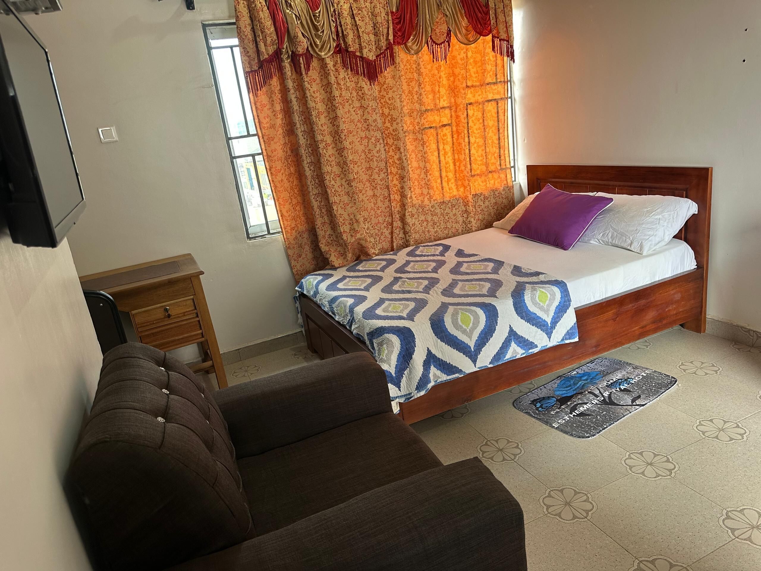 Une chambre à coucher comprenant un lit, une chaise, une petite table, une fenêtre avec des rideaux et un tapis à motifs.