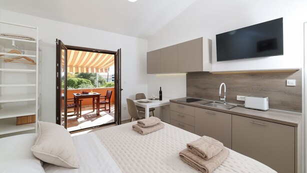 Quarto com cama, mesa de cabeceira, televisão montada acima da cama, área de cozinha com pia e torradeira, e porta de vidro deslizante que dá para uma área de estar externa.