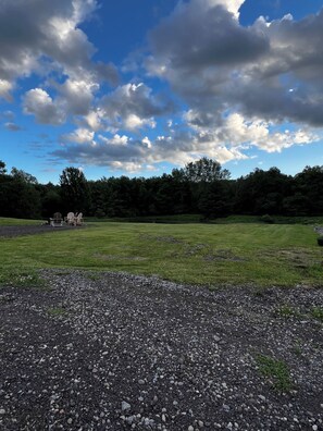 A gravel area with a grassy field, trees, and a cloudy sky.