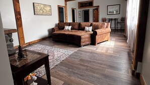 A spacious living area with a brown leather sofa, a coffee table, and a rug. There are wooden beams on the ceiling, and a dining area is visible in the background.