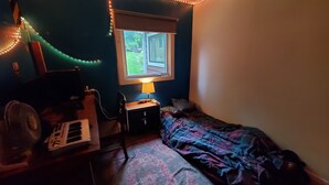 Une petite chambre à coucher avec un lit, un bureau avec une lampe, une chaise, un ventilateur et une fenêtre donnant sur l’extérieur.