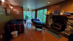 Un salon chaleureux avec une cheminée, un plancher en bois et de grandes fenêtres donnant sur un paysage verdoyant.