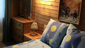 Ein Schlafzimmer mit Holzwand, einem Bett mit gemusterten Betttextilien, einem Nachttisch mit Lampe, einer Kommode und einem Gemälde.