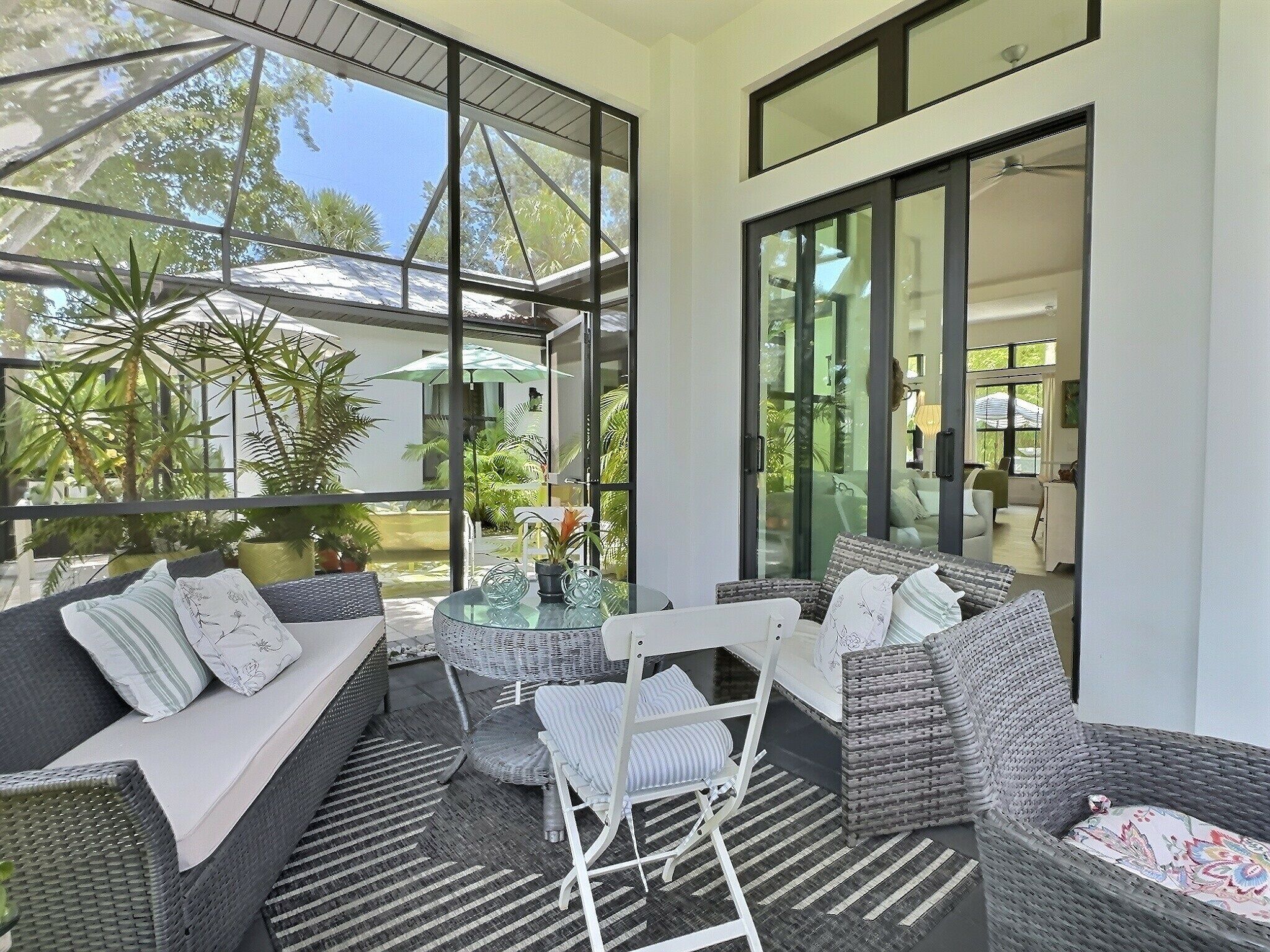 A modern outdoor living area with a sofa, chairs, and a glass table, surrounded by large windows and potted plants.