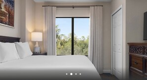 Ein Hotelzimmer mit einem großen Bett, einem Nachttisch mit Lampe, einem Kleiderschrank und Blick auf Grün durch das Fenster.