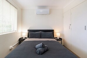 Ein Hotelzimmer mit Bett, zwei Nachttischlampen, einem Fenster mit Jalousien, einer Klimaanlage und einem Kleiderschrank.