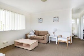 Ein Wohnzimmer mit beiger Couch, einem weißen Sessel, einem kleinen weißen Tisch und einem Holztisch. An den Wänden hängen zwei Bilder und es gibt ein Fenster mit Jalousien.