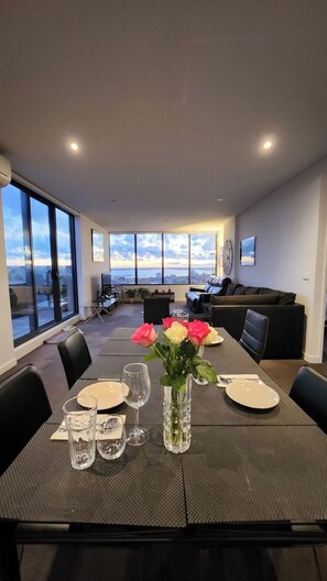 A dining area with a table set for four, large windows, a sofa, and a view of the cityscape. - Soak Above the City St Kilda Rooftop Jacuzzi Stay (St Kilda)