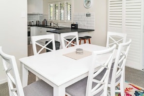A white dining table with four chairs, a kitchen in the background, and a clock on the wall.