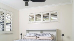 Une chambre à coucher comprenant un lit, deux tables de chevet, un ventilateur de plafond et une fenêtre avec des stores.