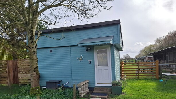 Un petit bâtiment bleu avec une porte blanche, entouré d’une cour verdoyante et d’une clôture en bois.