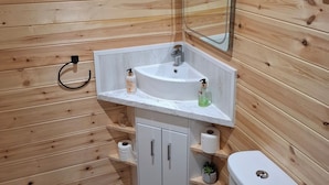 Une salle de bain aux murs lambrissés, avec un lavabo blanc, un miroir, des toilettes et des étagères.