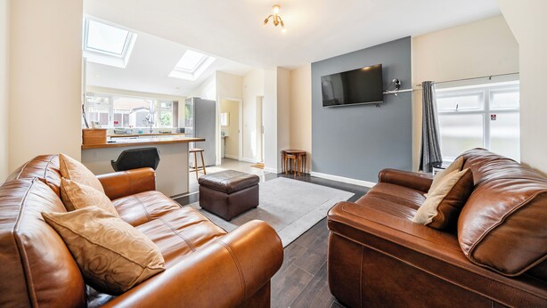 A modern living room with leather sofas, a flat-screen TV, and a kitchen in the background. - House Sleeps 4 - Free Parking and Garden (Manchester)