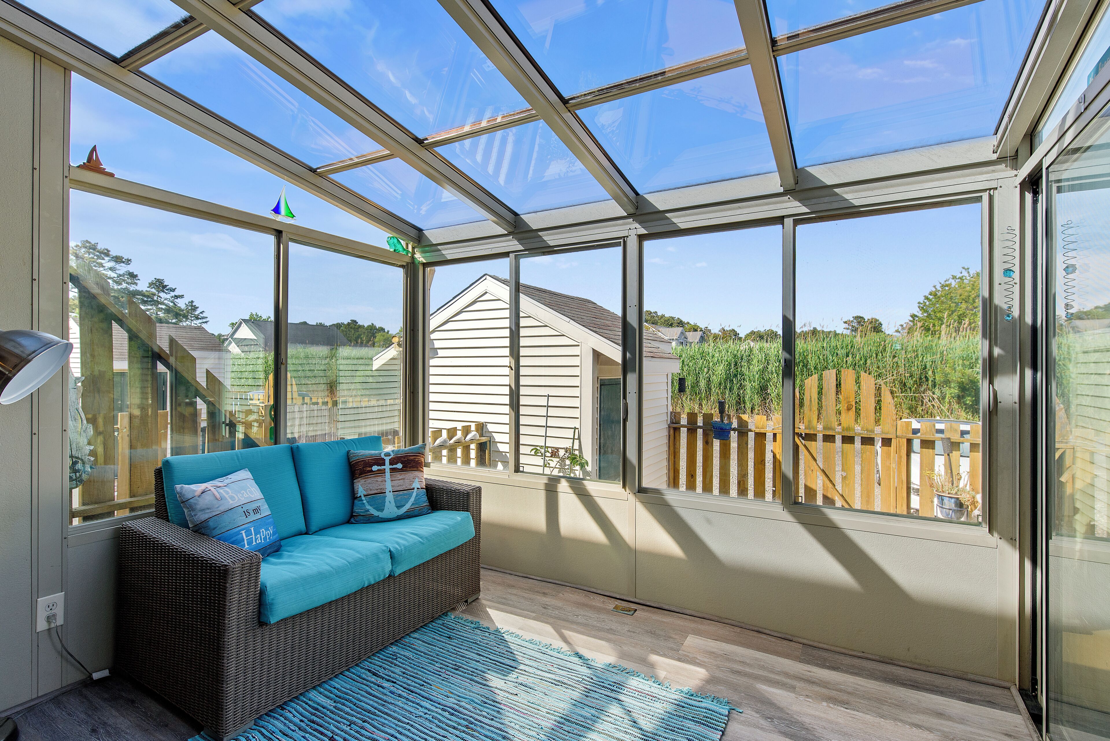 A sunroom with a glass ceiling, a wicker sofa with cushions, and a view of a wooden fence and house.
