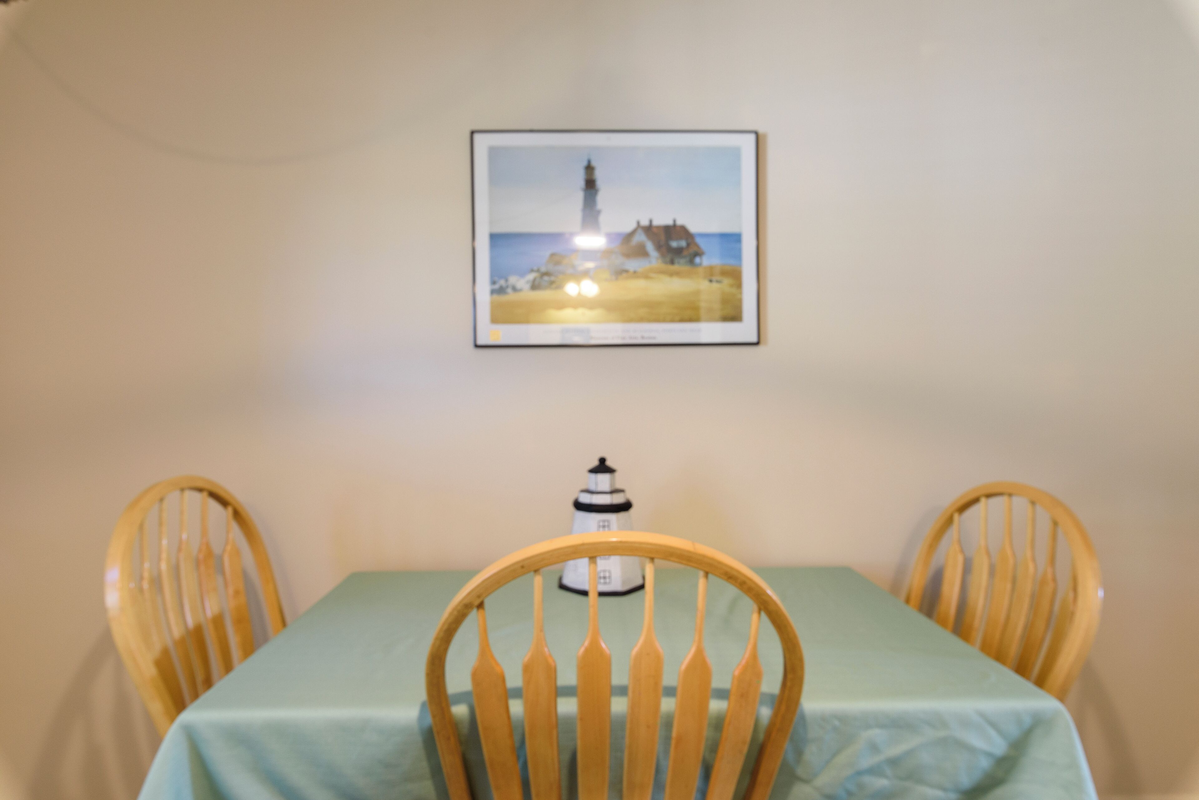 A dining table with a green tablecloth, two wooden chairs, and a framed picture of a lighthouse on the wall.