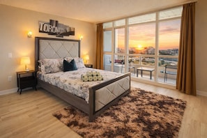 Un dormitorio con vista a la ciudad, una cama con ropa de cama estampada, dos lámparas de noche y un ventanal con cortinas.