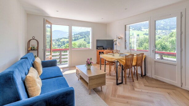 A spacious living area with a blue sofa, wooden dining table, and chairs, large windows with a view of greenery and hills. - L'Appartement des Sommets - Welkeys (Combloux)