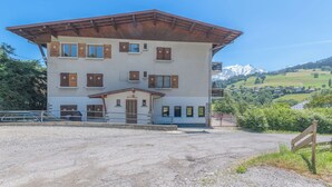 A multi-story building with a sloped roof, a gravel driveway, and a mountainous landscape in the background. - L'Appartement des Sommets - Welkeys (Combloux)