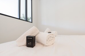 Ein ordentlich bezogenes Bett mit einem aufgerollten weißen Handtuch und einer schwarzen Willkommensbox.