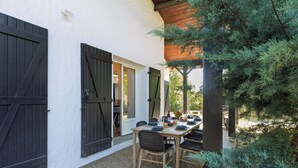 A patio with a table set for dining, surrounded by greenery and a building with large black shutters. - Villa Atlantide - Welkeys (Moliets-et-Maa)