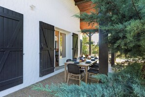 Une terrasse avec une table dressée pour le repas, entourée de verdure et bordée d’un bâtiment aux grandes volets noirs.