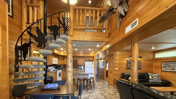 A wooden cabin interior with a spiral staircase, a dining area, a living room with a sofa, and a kitchen. - Family-friendly log cabin style house by Lewis & Clark Lake at DJ's Resort (Yankton)