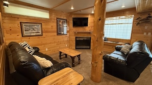 A log cabin interior with a fireplace, leather sofas, wooden tables, and a mounted television. - Family-friendly log cabin style house by Lewis & Clark Lake at DJ's Resort (Yankton)