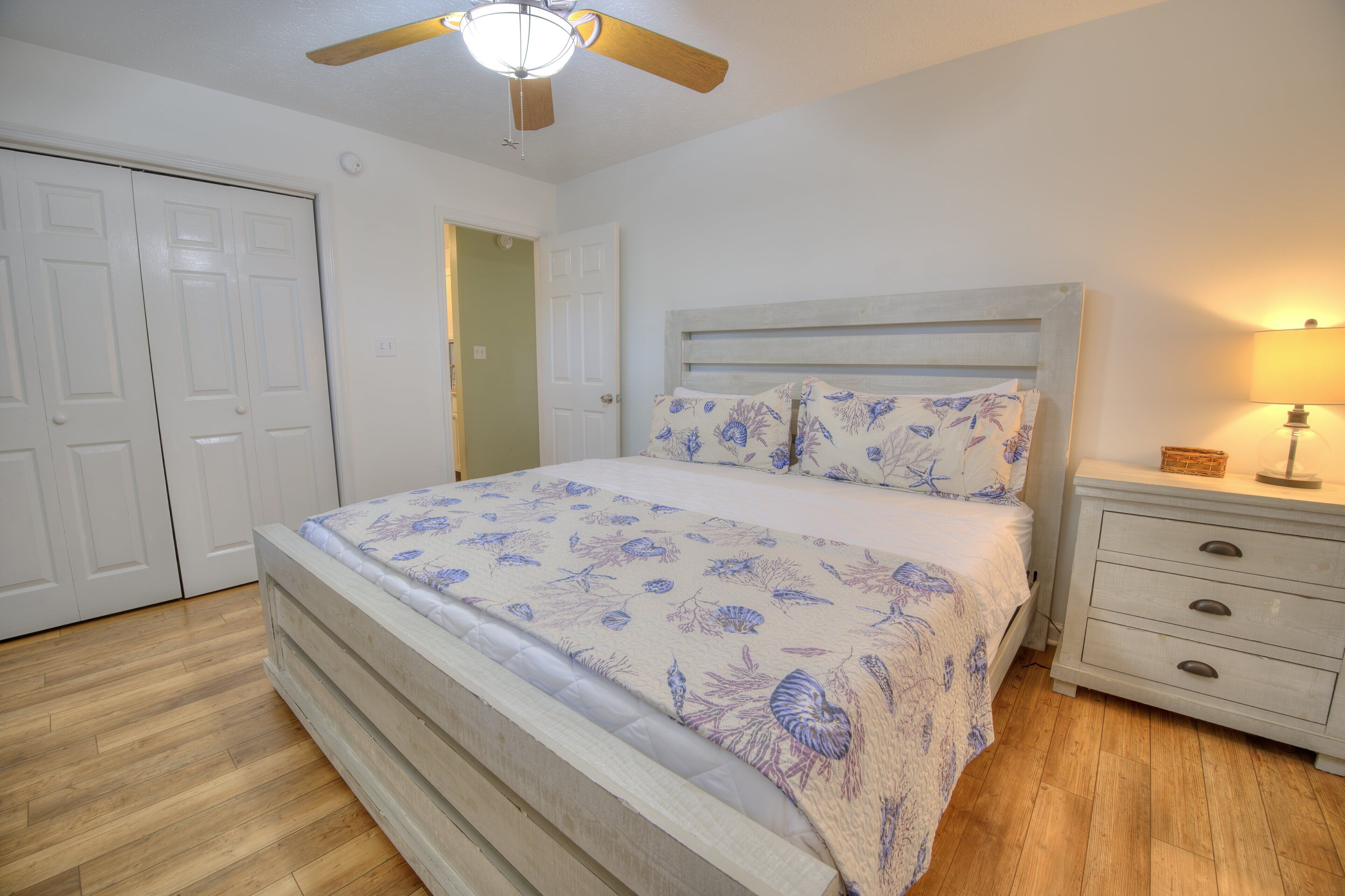 Une chambre à coucher comprenant un lit, une table de chevet, une lampe, un ventilateur de plafond et une porte.