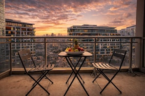 Ein Balkon mit zwei Stühlen, einem Tisch mit Obstschale und Blick auf die Stadt im Abendrot.