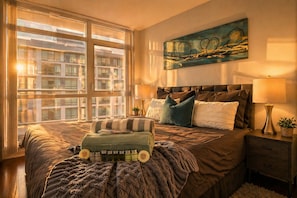 Ein Schlafzimmer mit großem Fenster, einem Bett mit vielen Kissen, einem Nachttisch mit Lampe und einem Bild an der Wand.