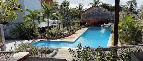 A swimming pool with a thatched roof structure, surrounded by tropical plants and a stone deck.