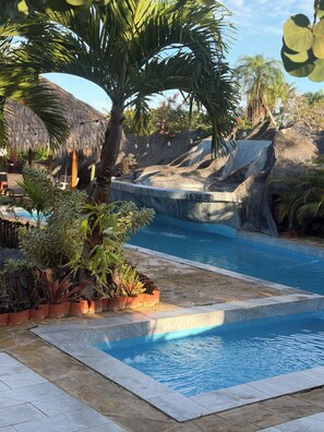 A swimming pool with a waterfall feature, surrounded by tropical plants and a thatched-roof structure.