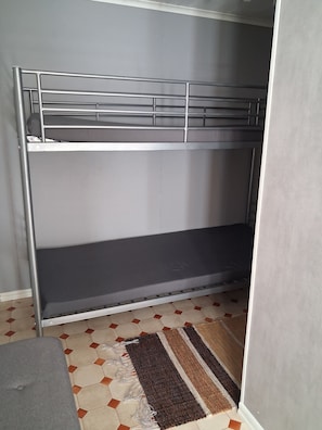 Una cama litera con barandillas metálicas, un colchón gris y una alfombra estampada sobre un suelo de baldosas.