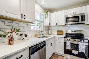Una cocina moderna con armarios blancos, electrodomésticos de acero inoxidable, lavavajillas y un ventanal con persianas.