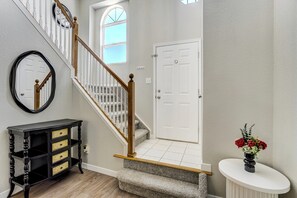 Una escalera con barandilla de madera, una puerta blanca, una consola negra con cajones y un jarrón blanco con flores.