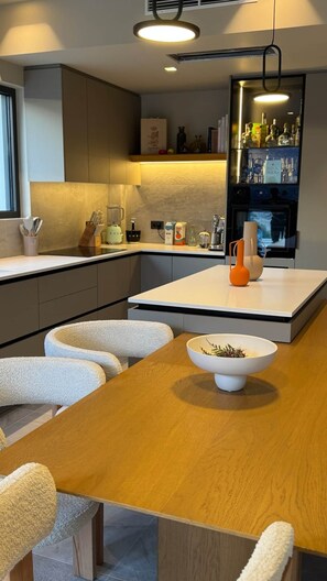 Eine moderne Küche mit Kochinsel, Essbereich mit Holztisch und weißen Stühlen sowie eine Bar mit Flaschen und Gläsern.
