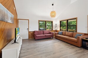 Ein großzügiges Wohnzimmer mit Holzboden, zwei Sofas, einem Fernseher und großen Fenstern mit Blick auf Bäume.