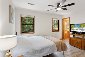 Ein Schlafzimmer mit Bett, Deckenventilator, Fernseher und zwei Fenstern.