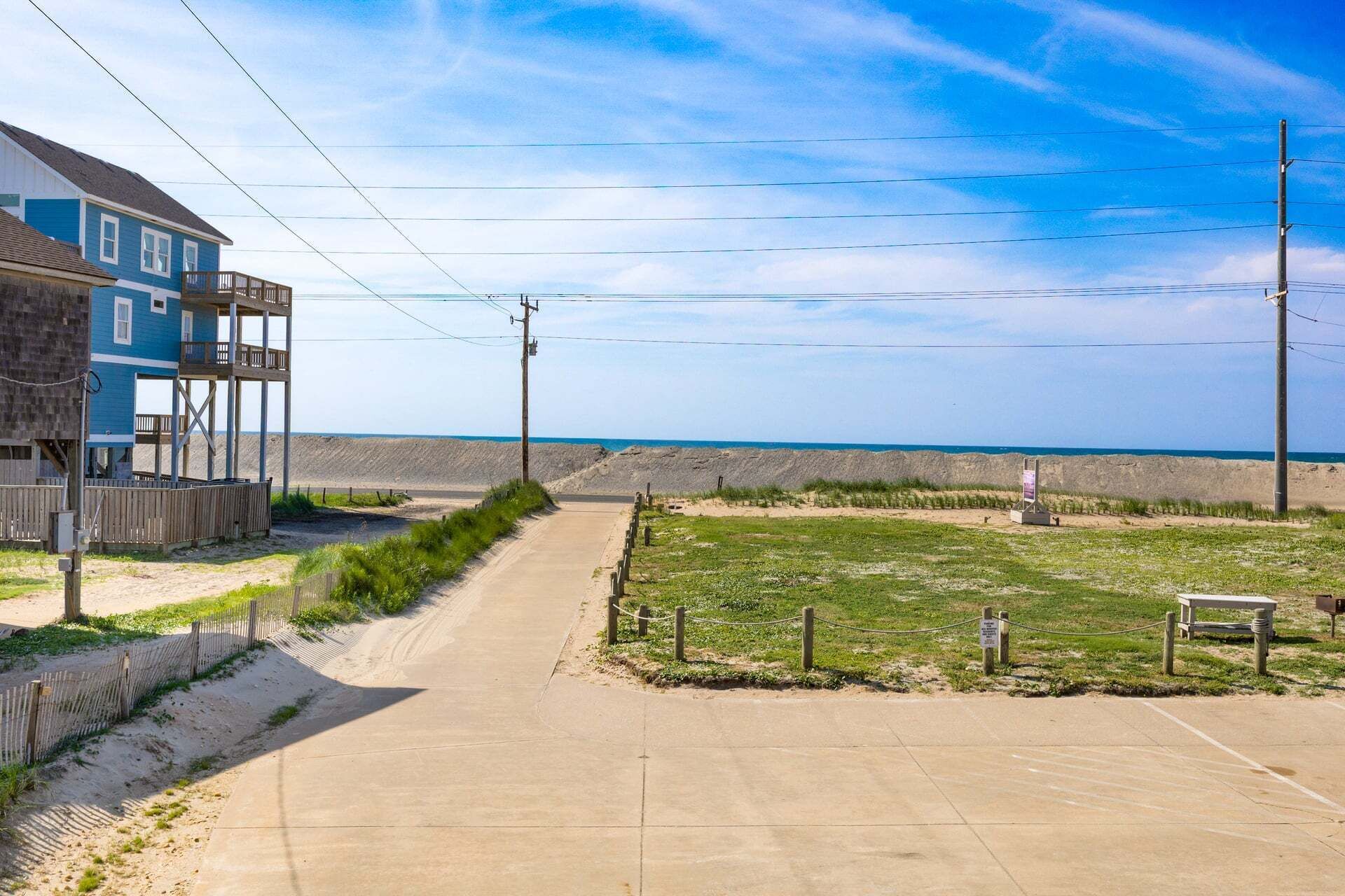 Een strandpand met een betonnen pad dat uitkomt op een zandgebied, een houten omheining en een gebouw met een balkon.
