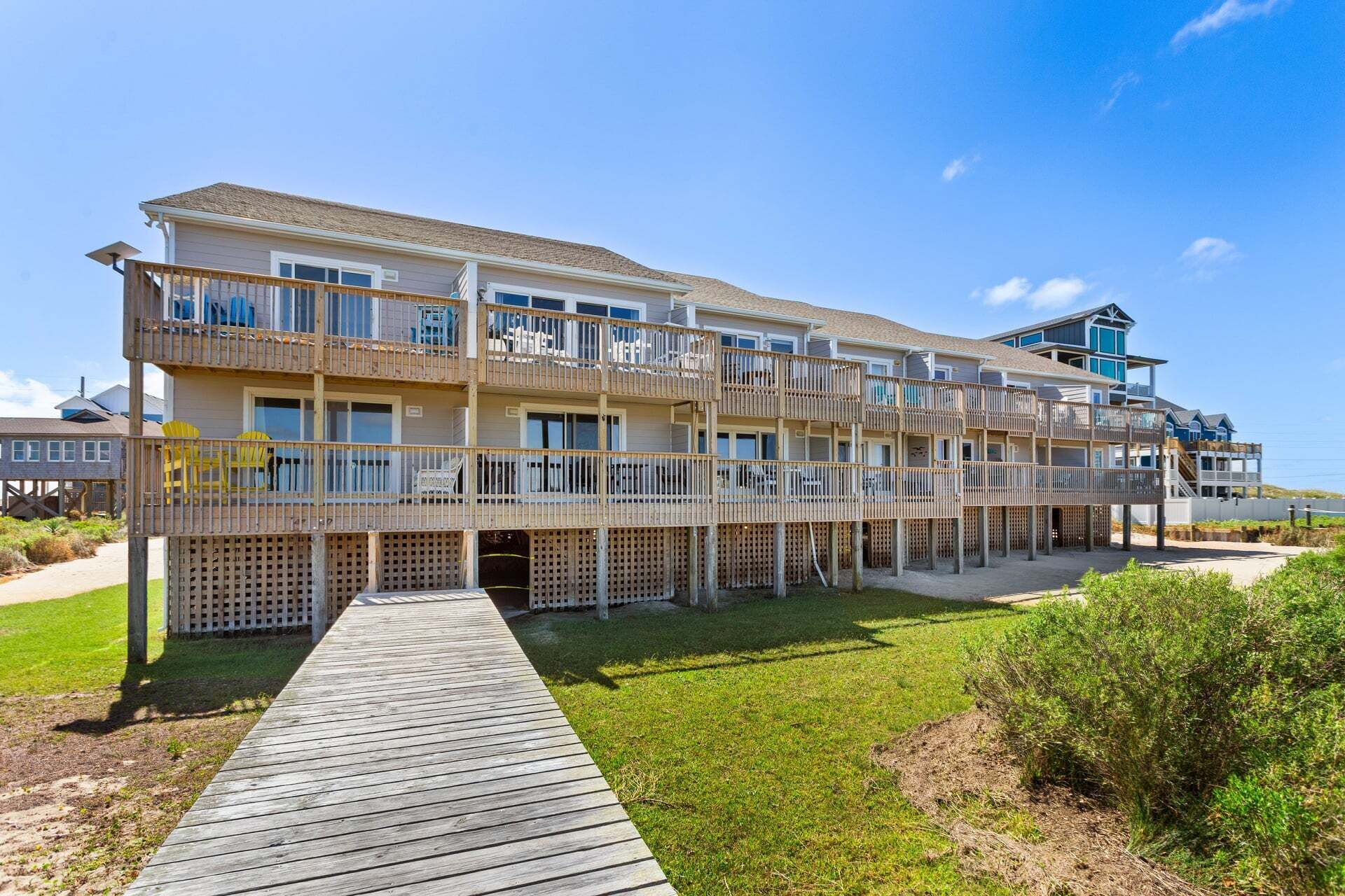 Un immeuble à plusieurs étages situé en bord de mer, doté de balcons, d’une terrasse en bois et surplombant un ciel dégagé.